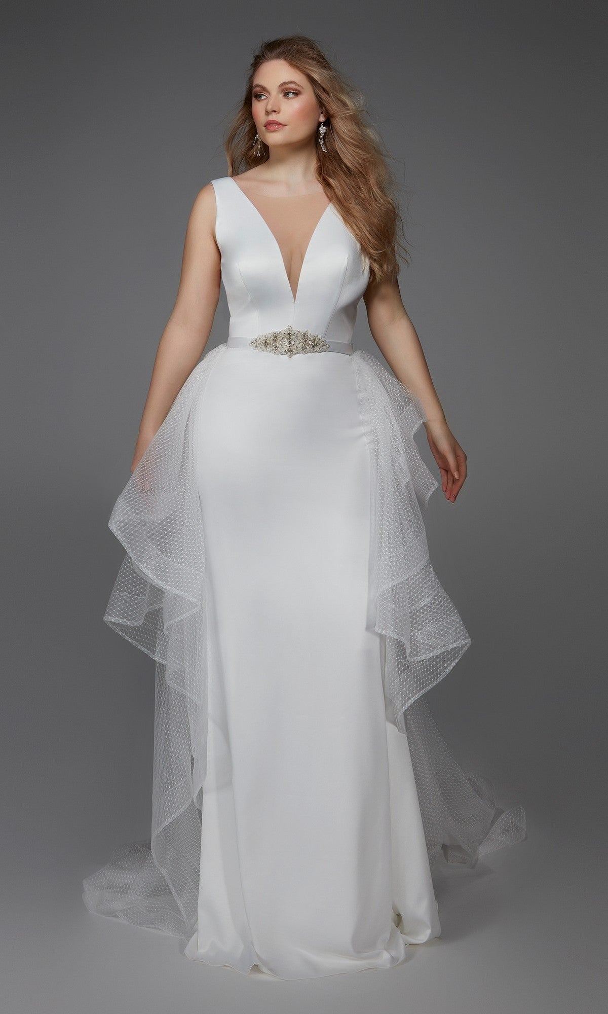 A woman stands against a plain gray background wearing the Alyce 7019—an elegant long dress in diamond white with a sleeveless deep V-neckline, embellished waist, and layered tulle overskirt.